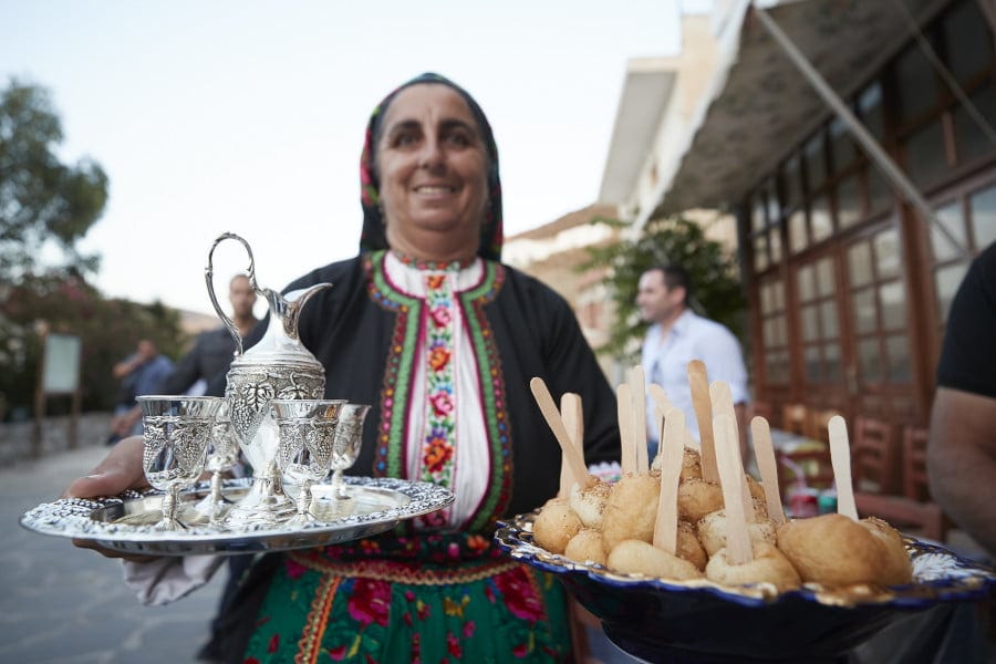 Karpathos, Lakki, Ai Giannis Festival - Gastronomy Tours woman dressed with folklore cloths smiling and holding plateau with sweets and glasses walking at monastery of Ai Giannis area, in Lakki, Greece that commemorate festival of her birth